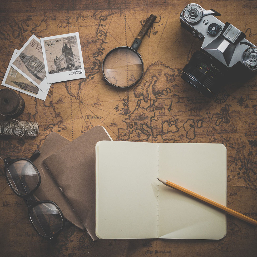 A collection of travel-themed items laid out on an old sepia map. Open notebook and pencil are predominantly featured.