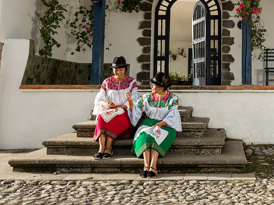 Two women in traditional Andean clothing embroidering on stone steps, capturing cultural moments on a Machu Picchu trip.