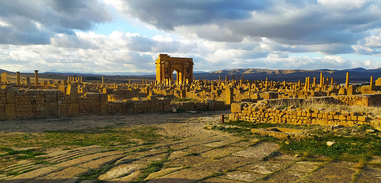 Ancient ruins at a UNESCO site in Algeria with stone structures and mountains under a cloudy sky.