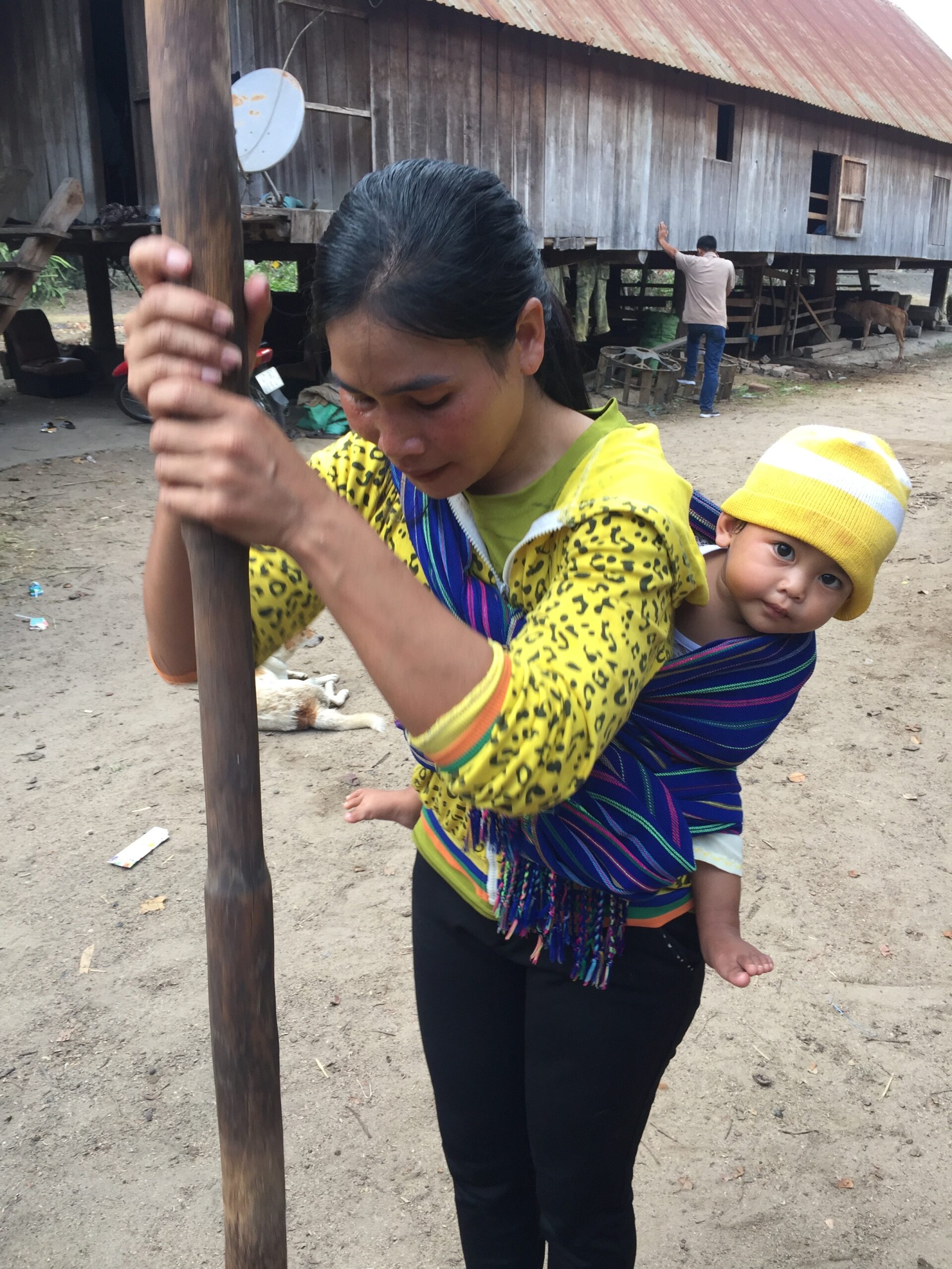 Travel Photo: Woman grinding coffee beans with a traditional pole while a baby is strapped to her back in Vietnam.